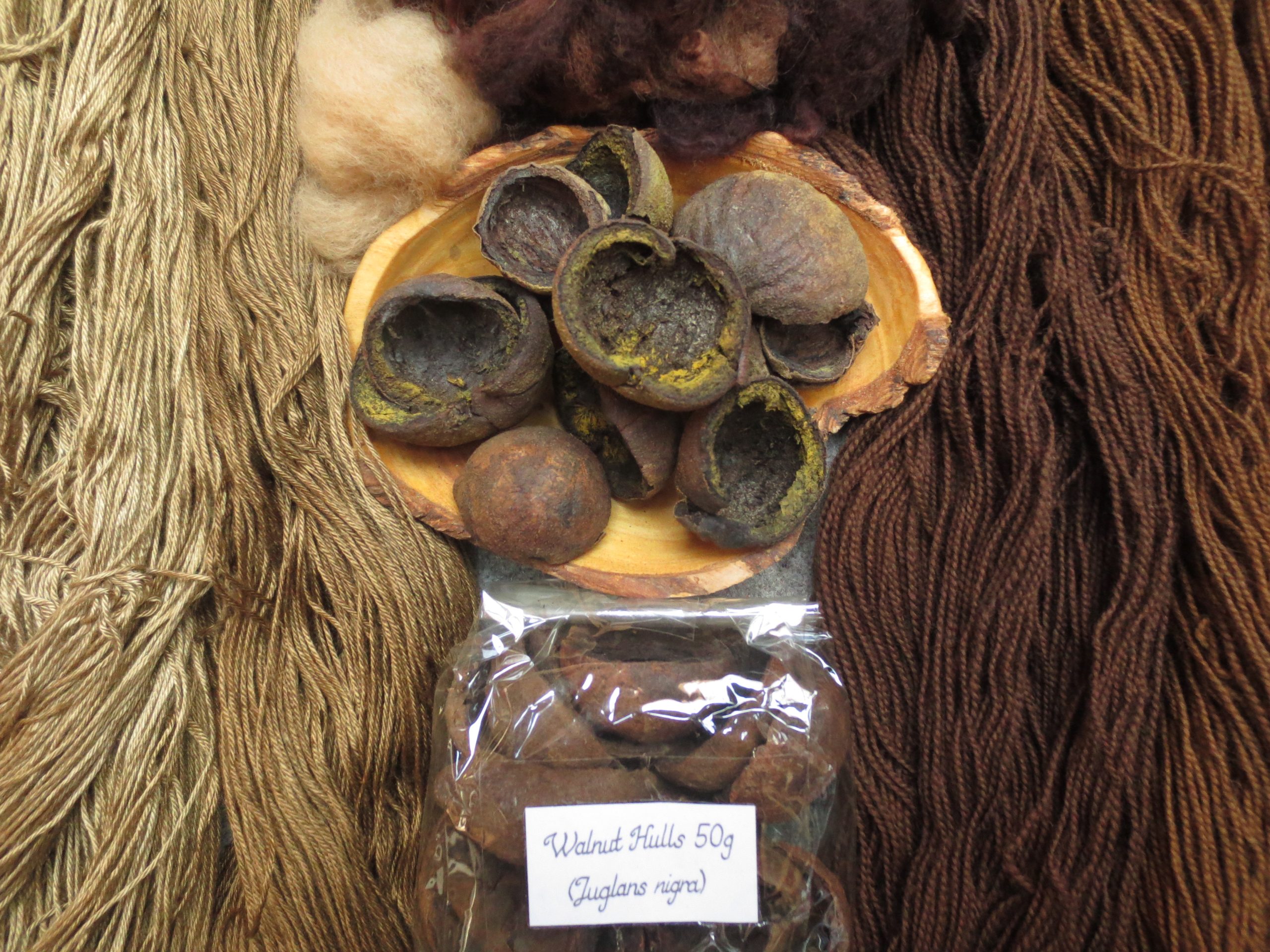 Wooden bowl of walnut hulls surrounded by wool and silk samples in shades of brown.