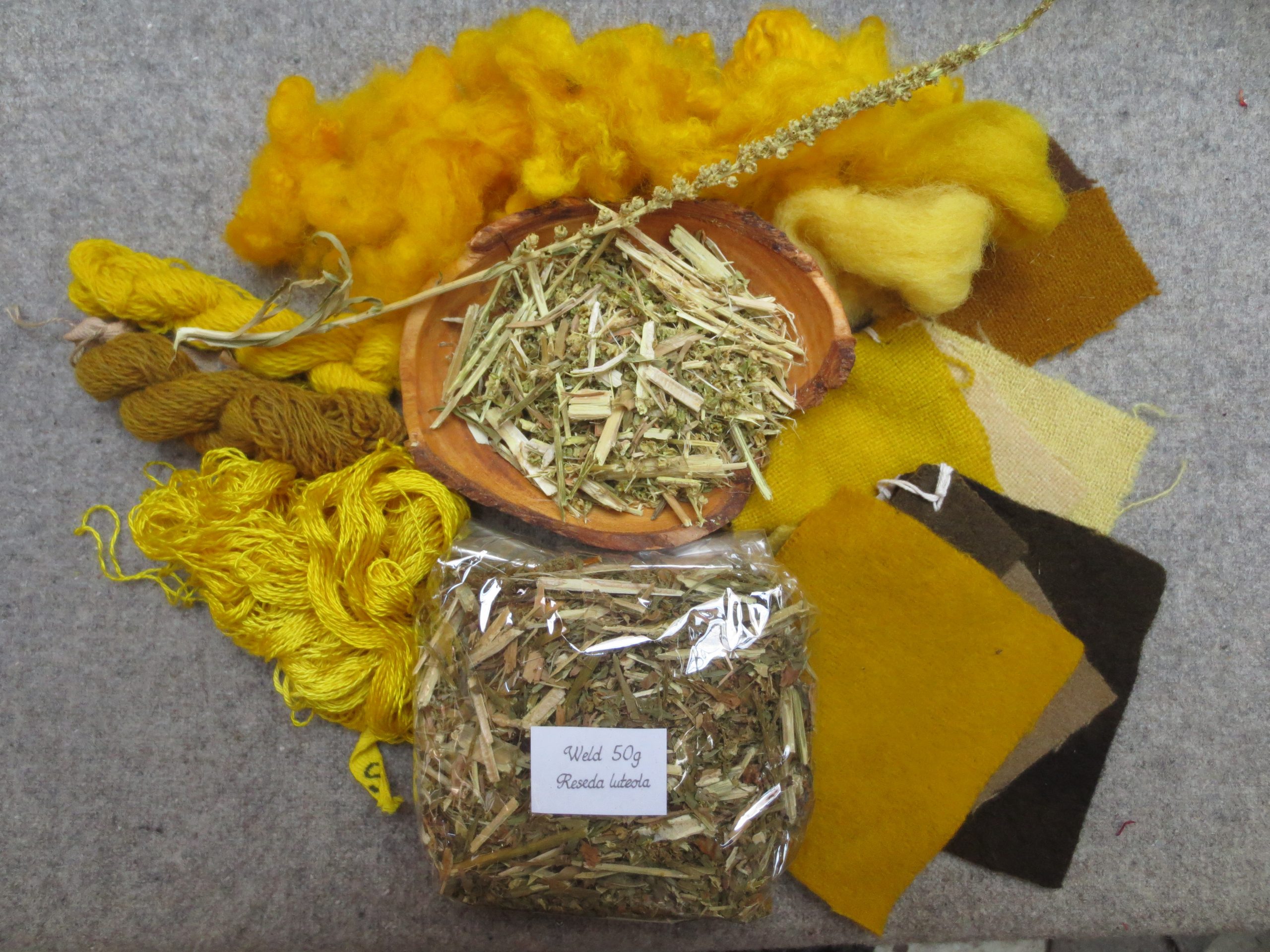 Wooden bowl of dried weld surrounded by yellow and grey, brown wool and silk samples.
